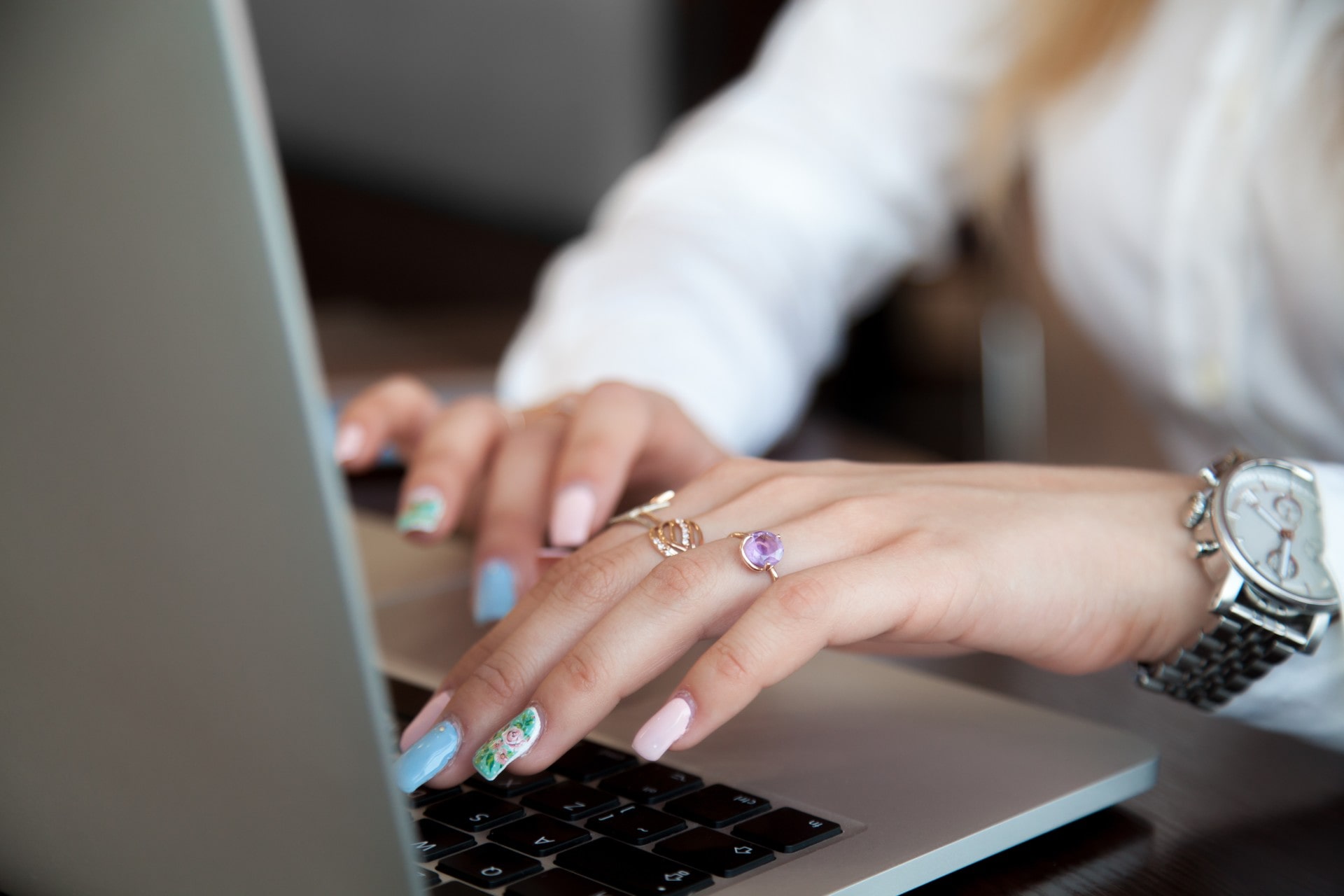 A close-up of a woman&rsquo;s hands typing on a computer, her nails are colorful in design, and she is wearing a silver watch.
