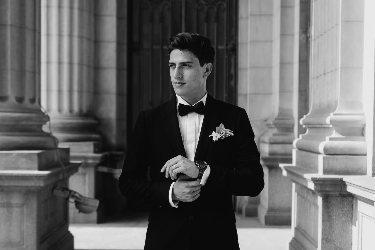 A black and white image of a man in a tuxedo surrounded by large pillars, adjusting his cuff and showcasing his watch.