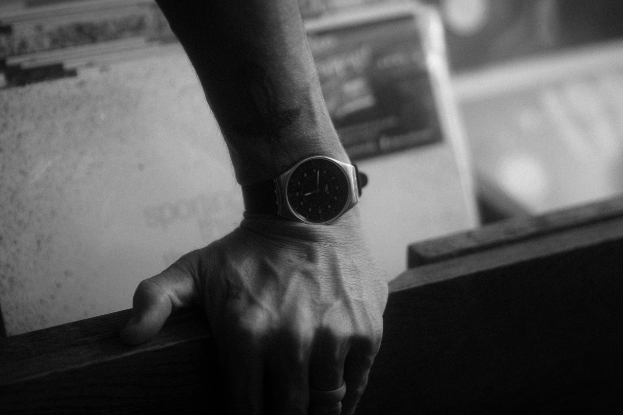 A black and white image of a man&rsquo;s hand resting on a wooden surface, his wrist showcasing an elegant watch.