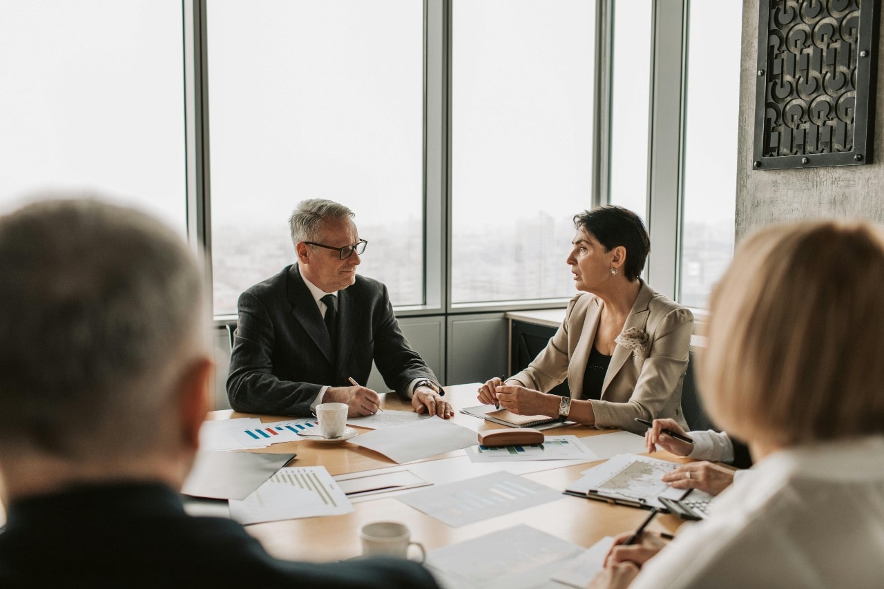 A group of business professionals sat around a table together, the table covered in papers and coffee mugs.