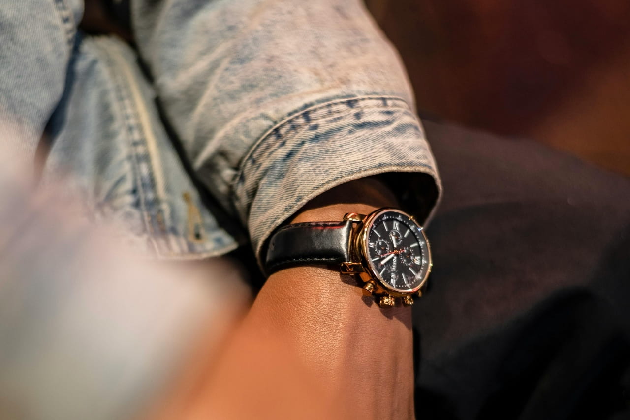 A person in a denim jacket wearing a luxury watch with a black leather strap, a yellow gold bezel, and a black dial.