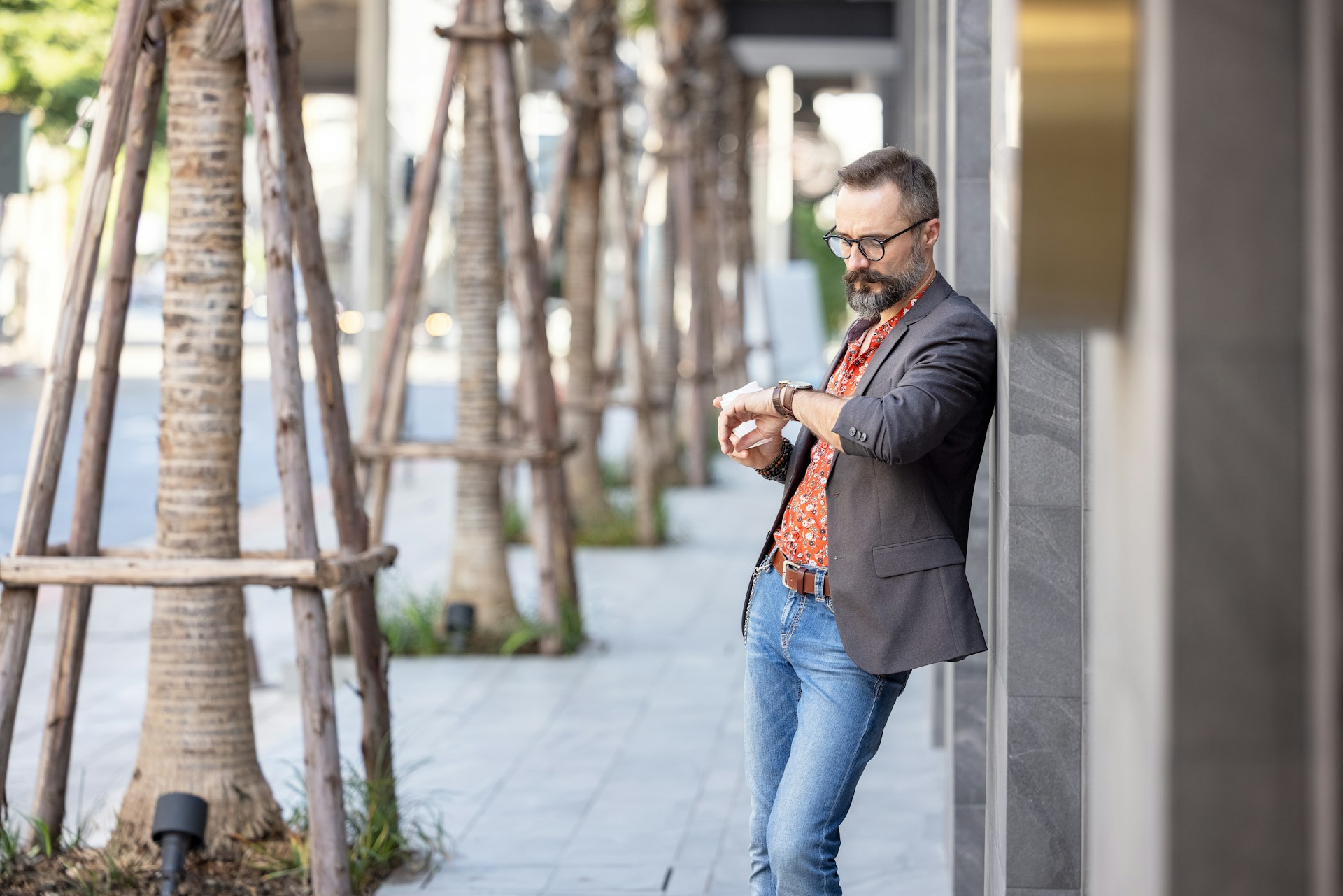 A man in a business casual outfit rests against a building and checks his watch.