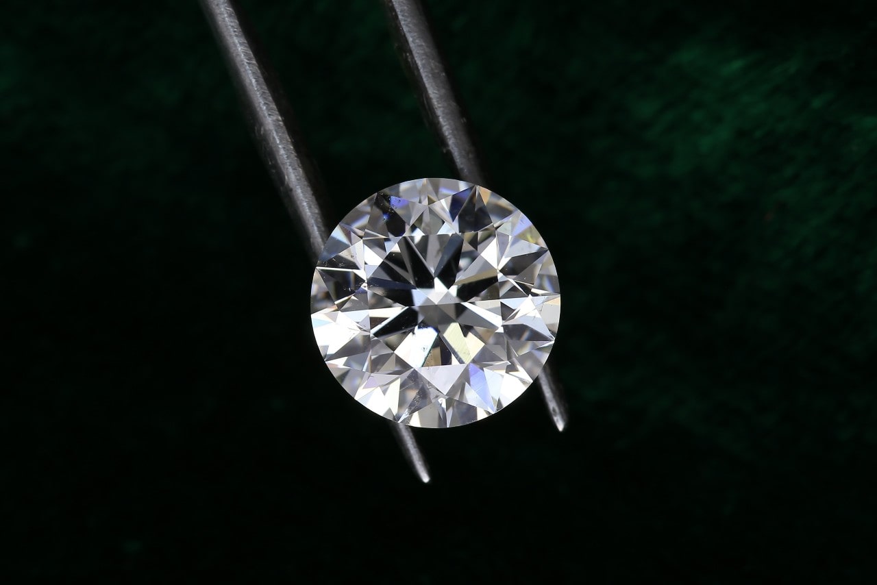 A close-up of a round diamond held by a pair of tweezers on a black background.