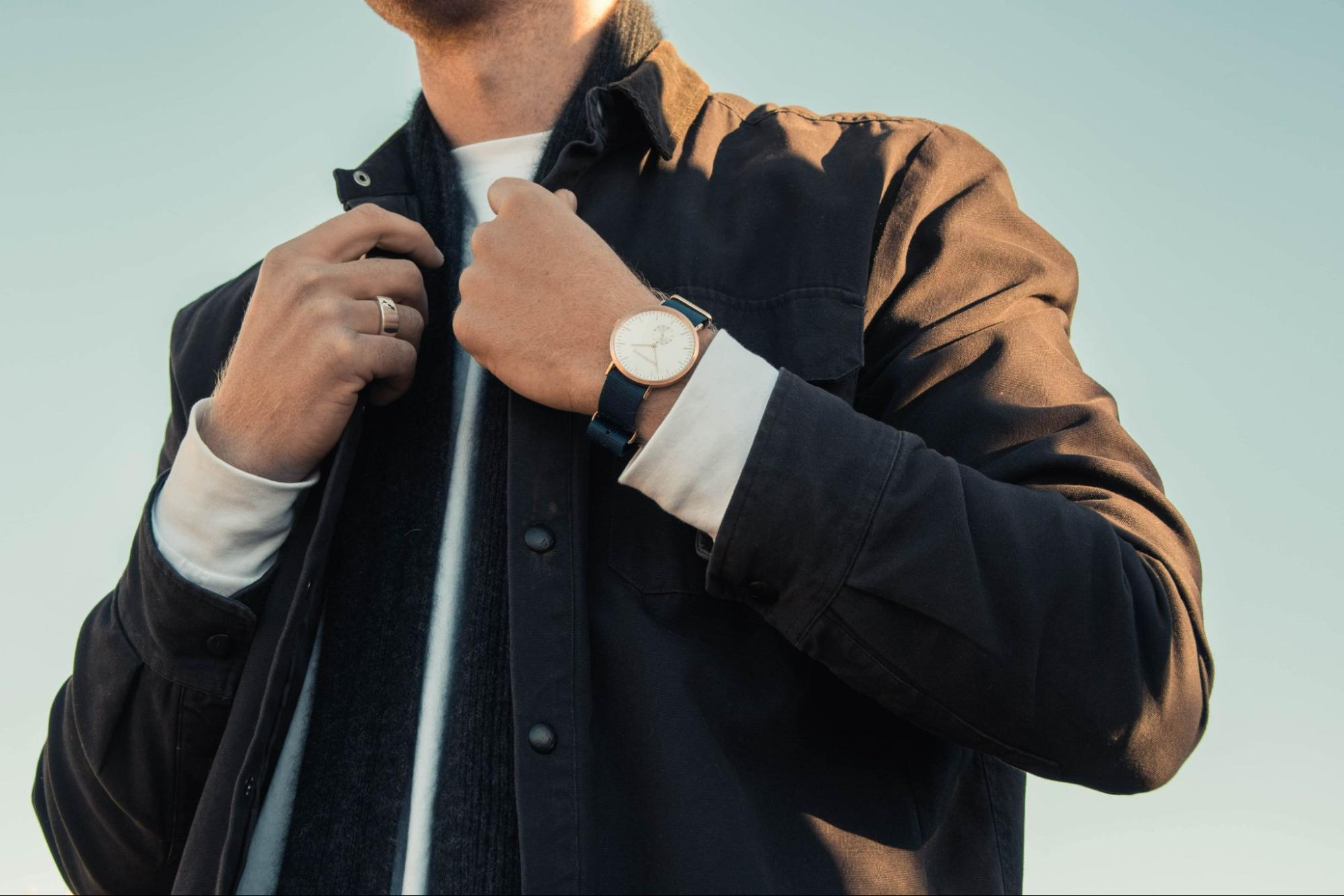 A man touches his coat’s collar, wearing a minimalist watch.