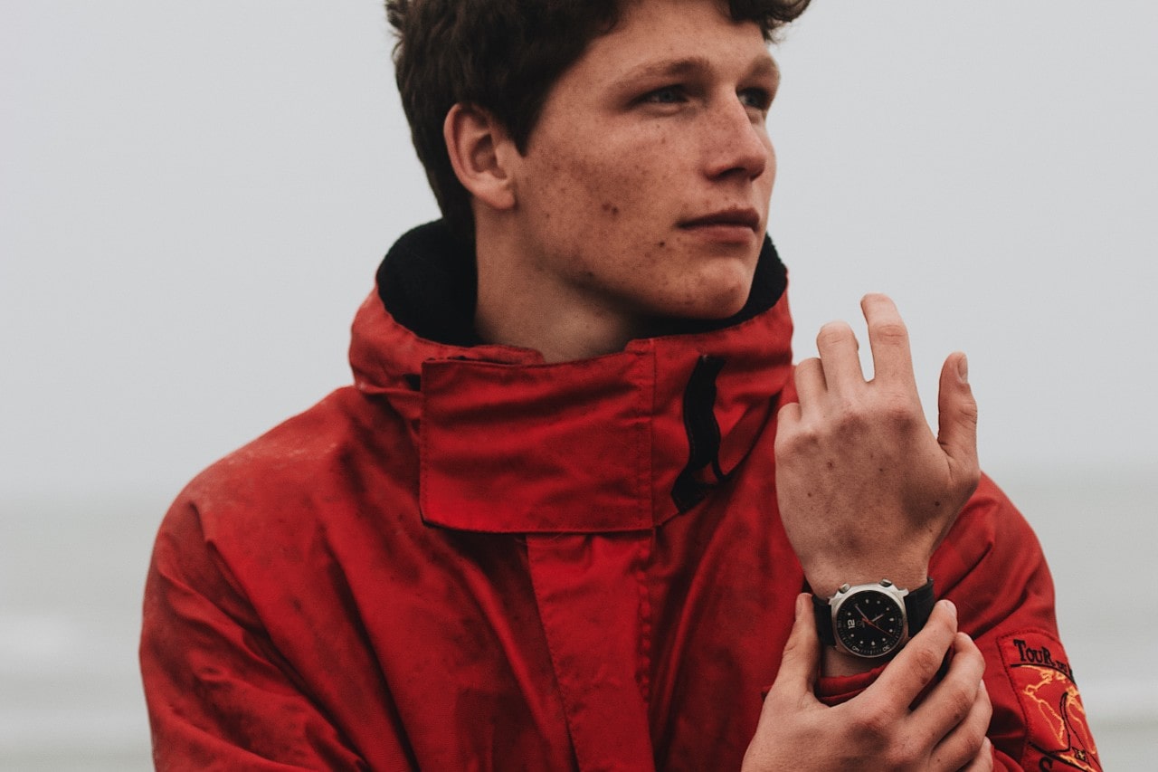 A man in a red windbreaker jacket holding his wrist, showcasing his silver watch with a black watchface.