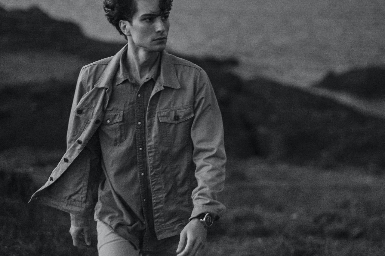 A black and white image of a man walking in an open field with water in the distance, his shirt blowing in the wind and his watch glimmering in the sun.