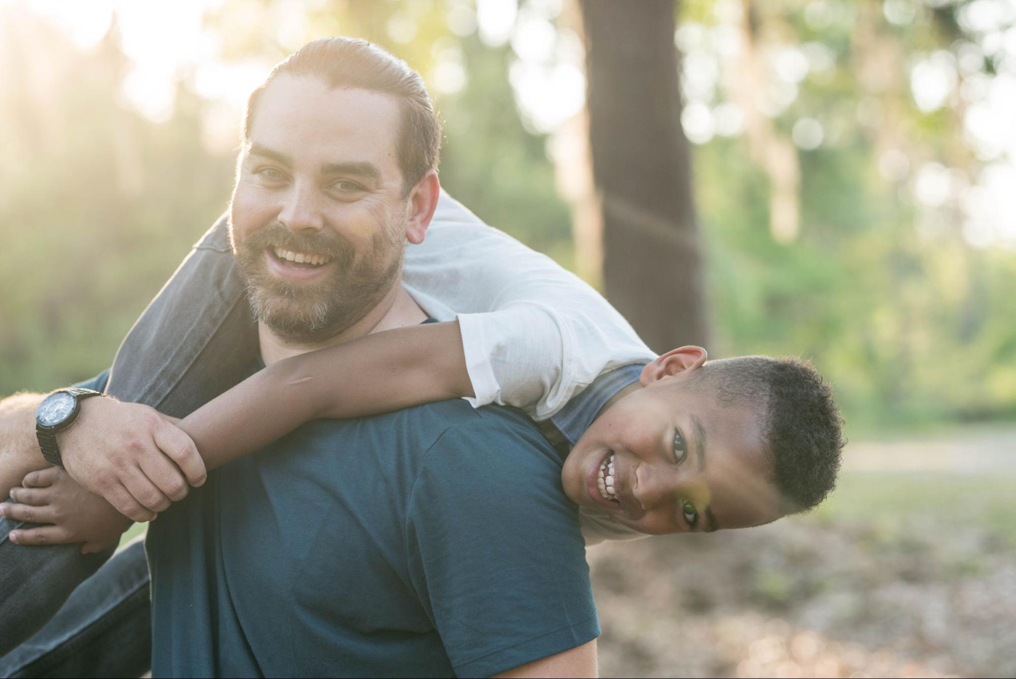 A smiling father carries a young child on his shoulders in a sunlit park.