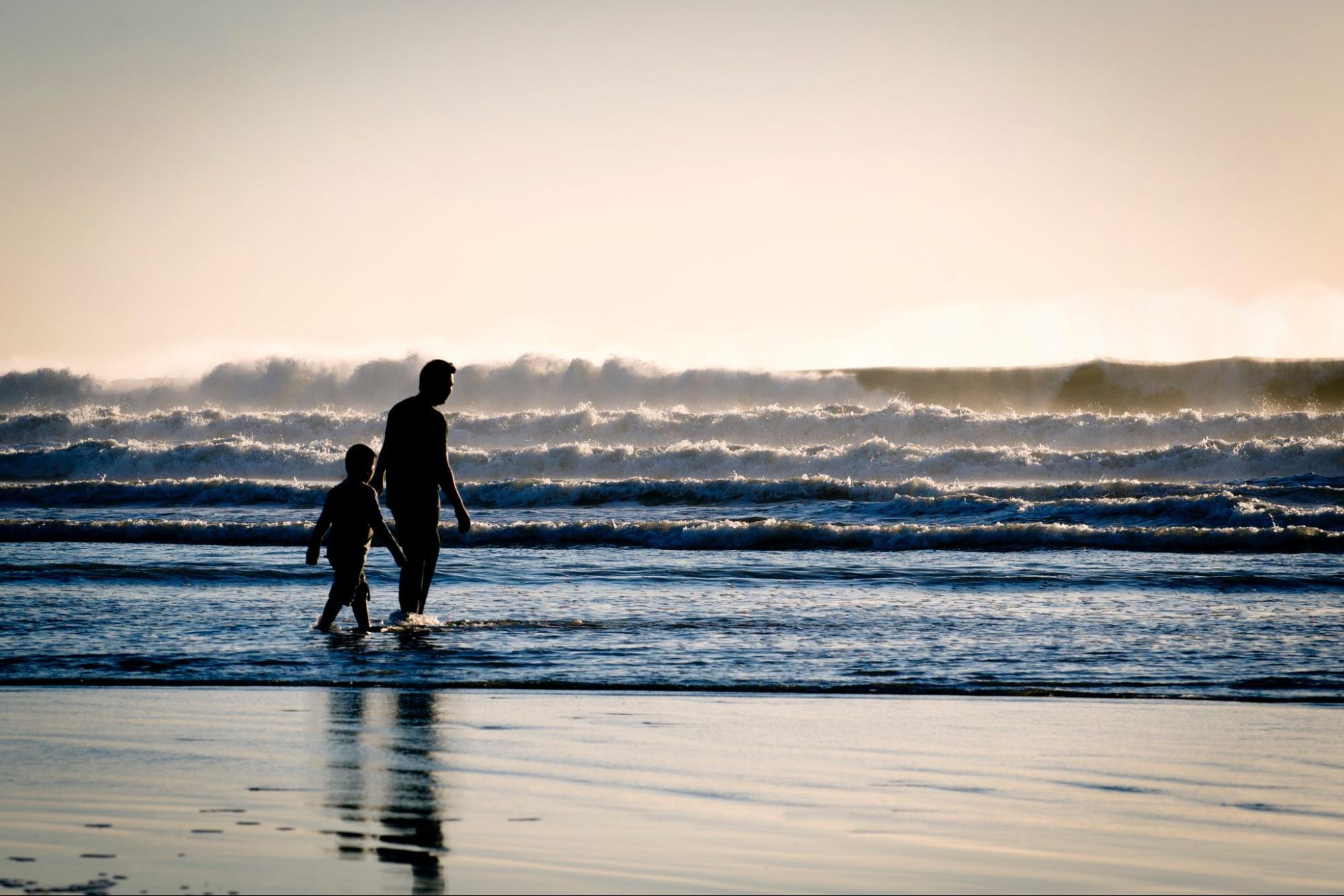 Silhouettes of an adult and child walk together along the shoreline at sunset.