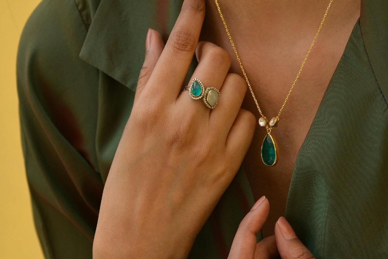 A woman’s hand raised to her chest, wearing a gemstone toi et moi ring and a gemstone necklace.