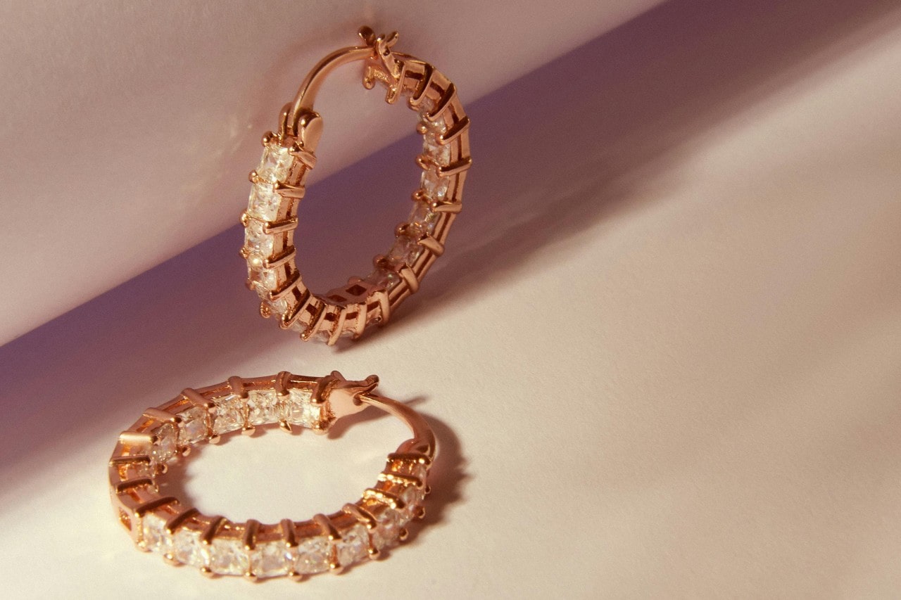 A pair of yellow gold princess cut diamond hoop earrings on a beige surface.