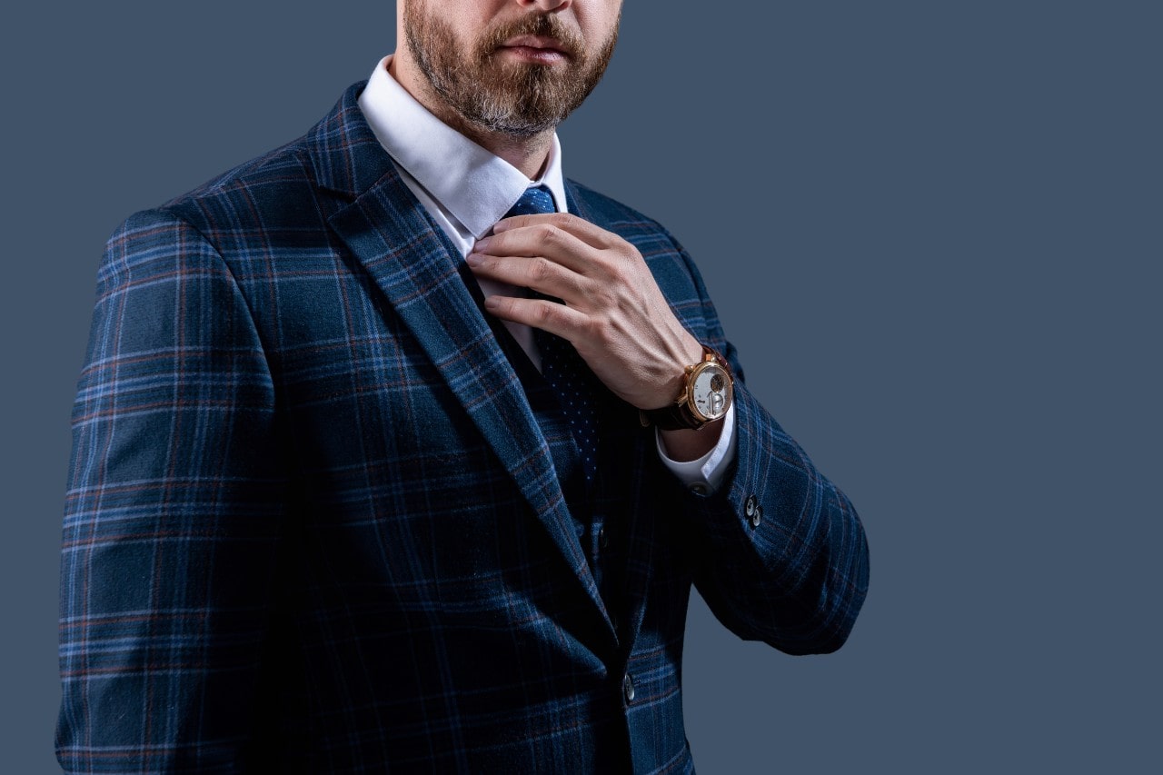 A man adjusts his tie while wearing a blue plaid suit and a watch.