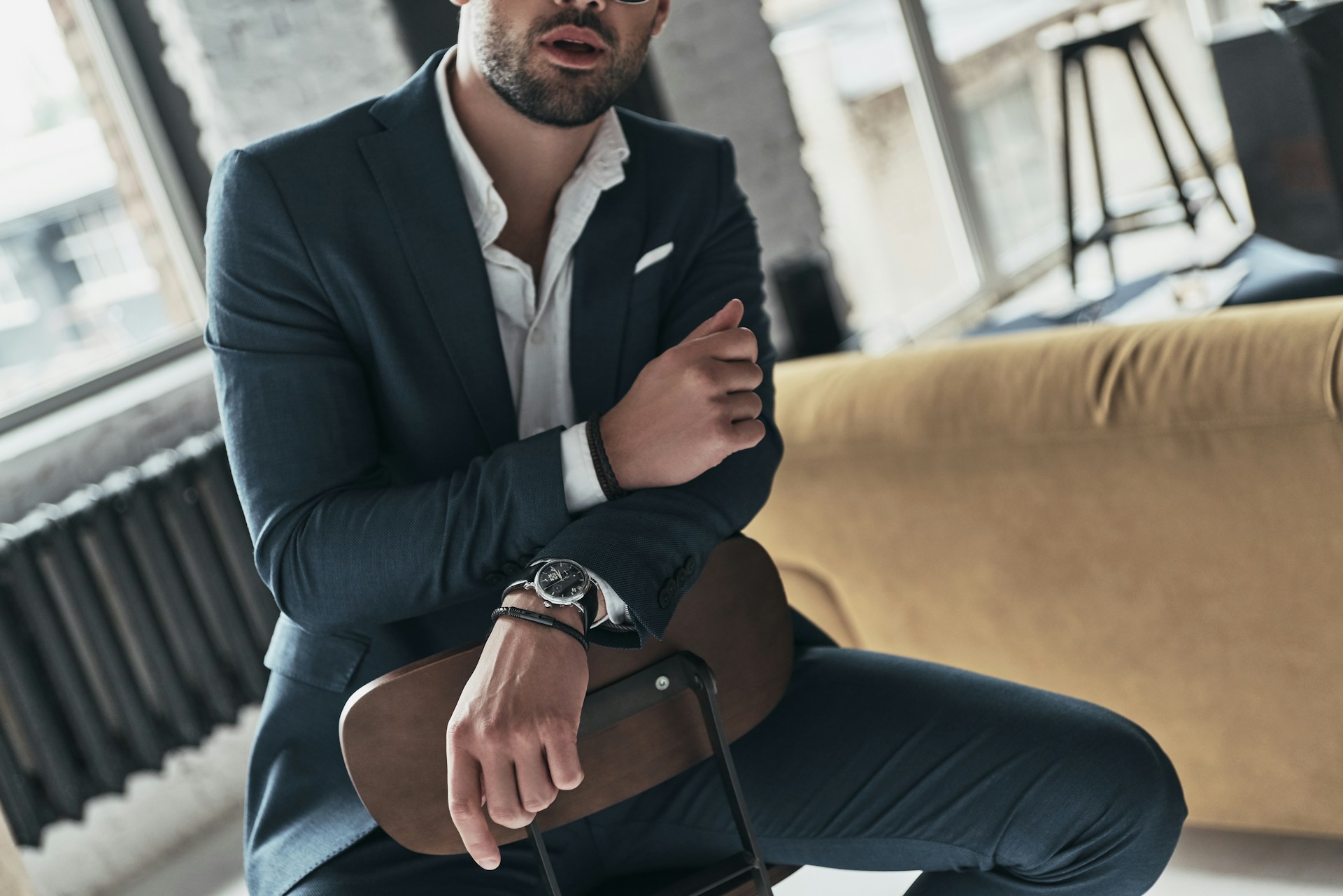 A man in a dark suit wearing a white shirt, wristwatch, and sunglasses sits casually on a chair indoors, leaning slightly.