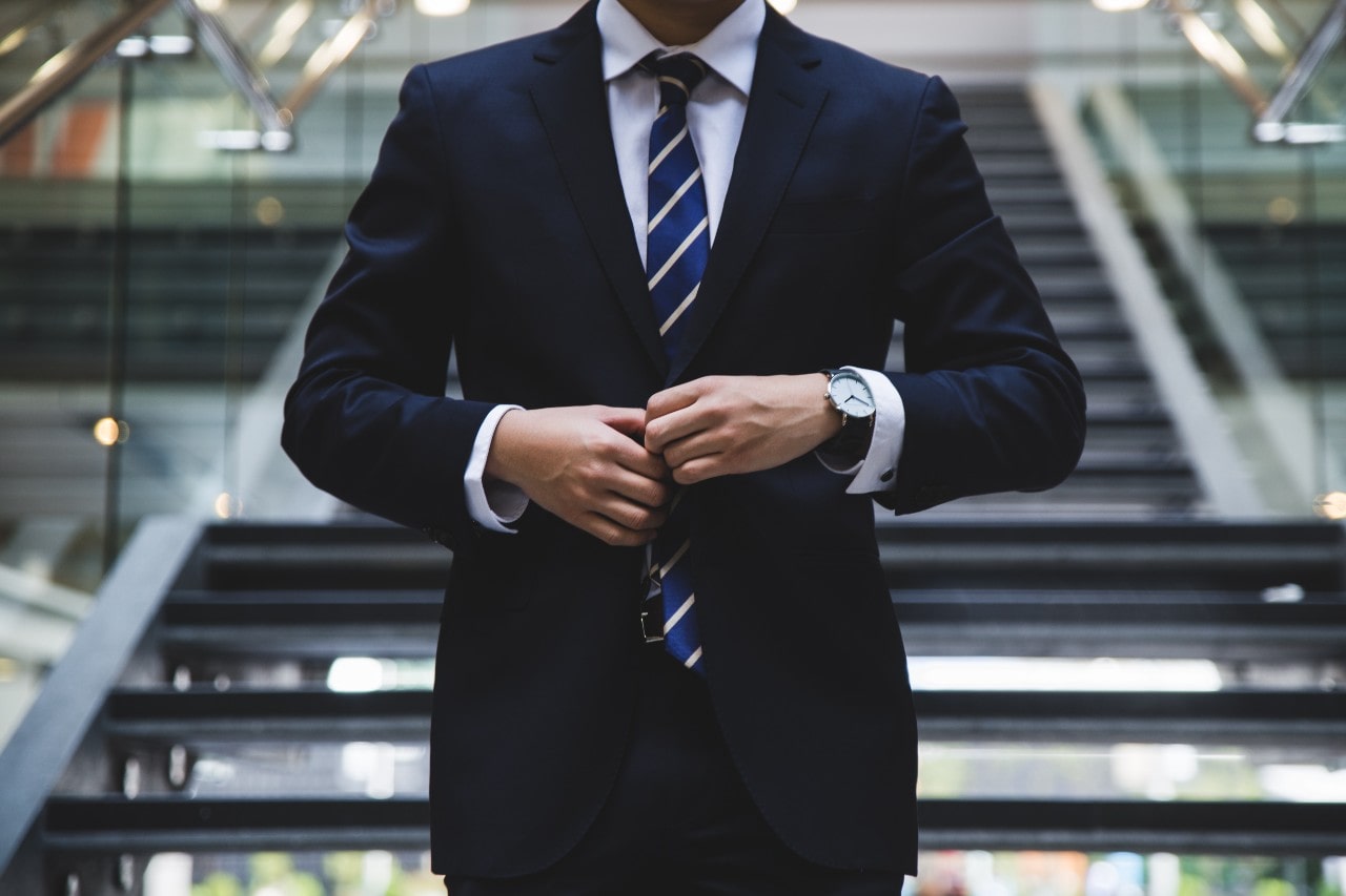 A man in a dark suit wearing an elegant watch adjusts his jacket, standing confidently on a modern staircase.