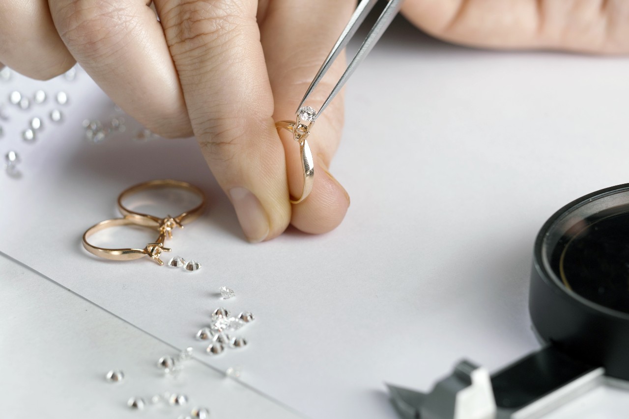 Close up of jeweler’s hand using tweezers to set a diamond on a gold ring amid scattered loose diamonds. Nearby are two more rings and a magnifying glass Close up of jeweler’s hand using tweezers to set a diamond on a gold ring amid scattered loose diamonds. Nearby are two more rings and a magnifying glass