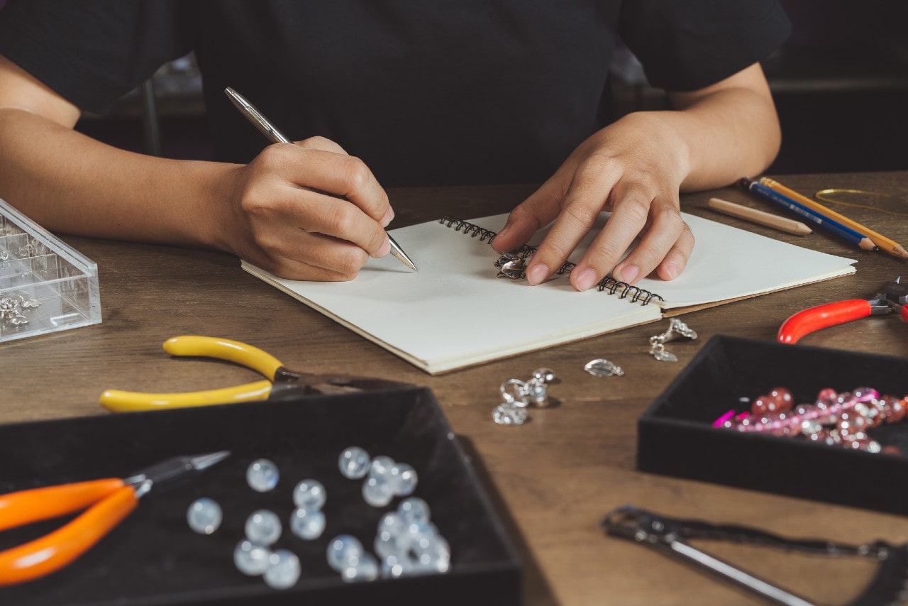 Close up of jeweler’s hands sketching a jewelry design in a notebook on a wooden table, surrounded by tools, beads, and gemstones. Close up of jeweler’s hands sketching a jewelry design in a notebook on a wooden table, surrounded by tools, beads, and gemstones.