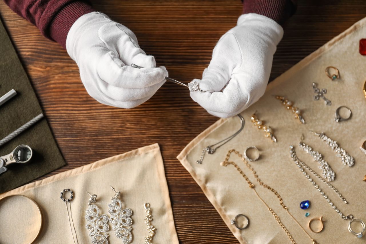 A jeweler wearing white gloves examines a diamond pendant with tweezers with various jewelry pieces assorted on beige clothes A jeweler wearing white gloves examines a diamond pendant with tweezers with various jewelry pieces assorted on beige clothes