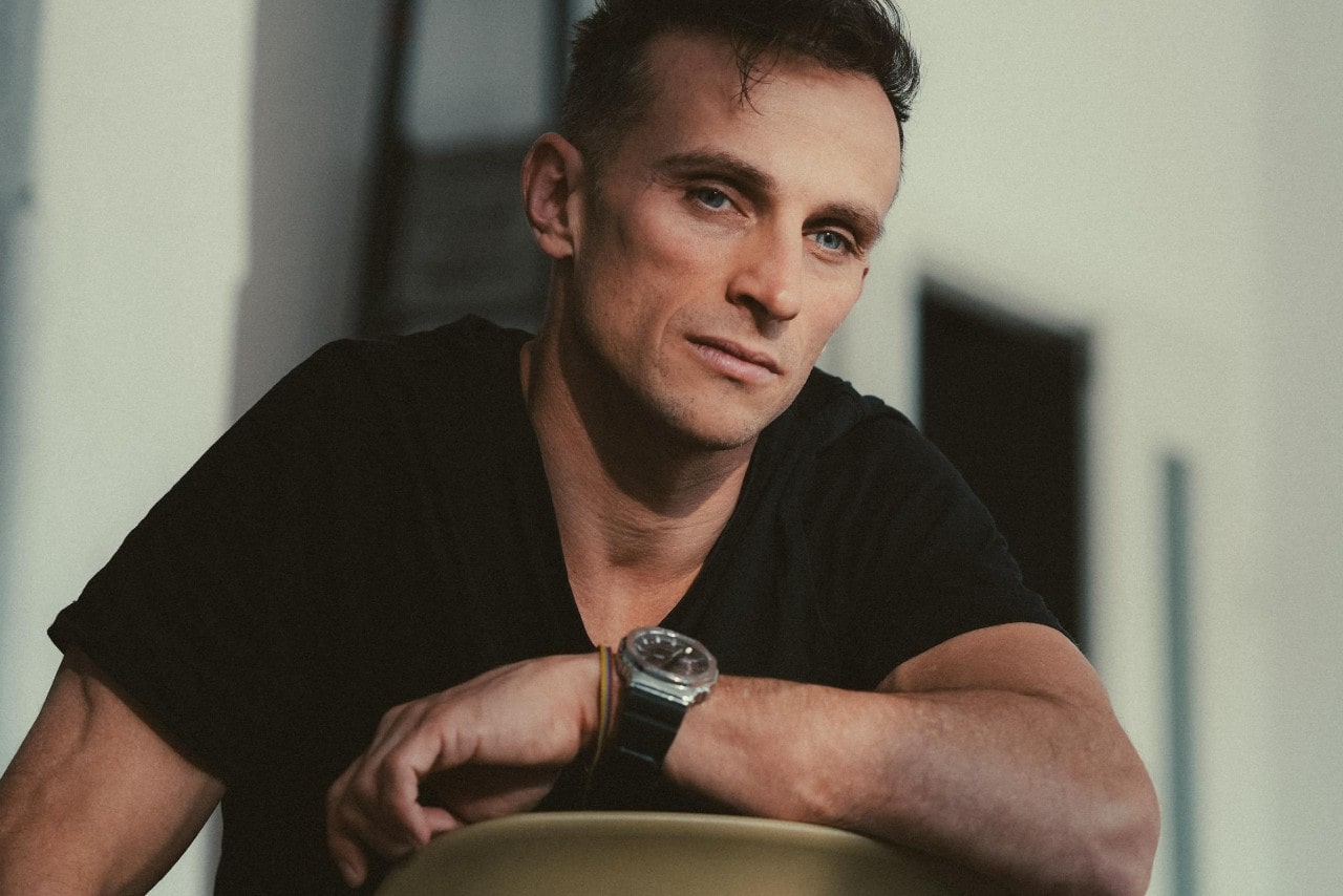 A man in a black T-shirt wearing a luxury wrist watch leans on a chair, looking thoughtful. A man in a black T-shirt wearing a luxury wrist watch leans on a chair, looking thoughtful.