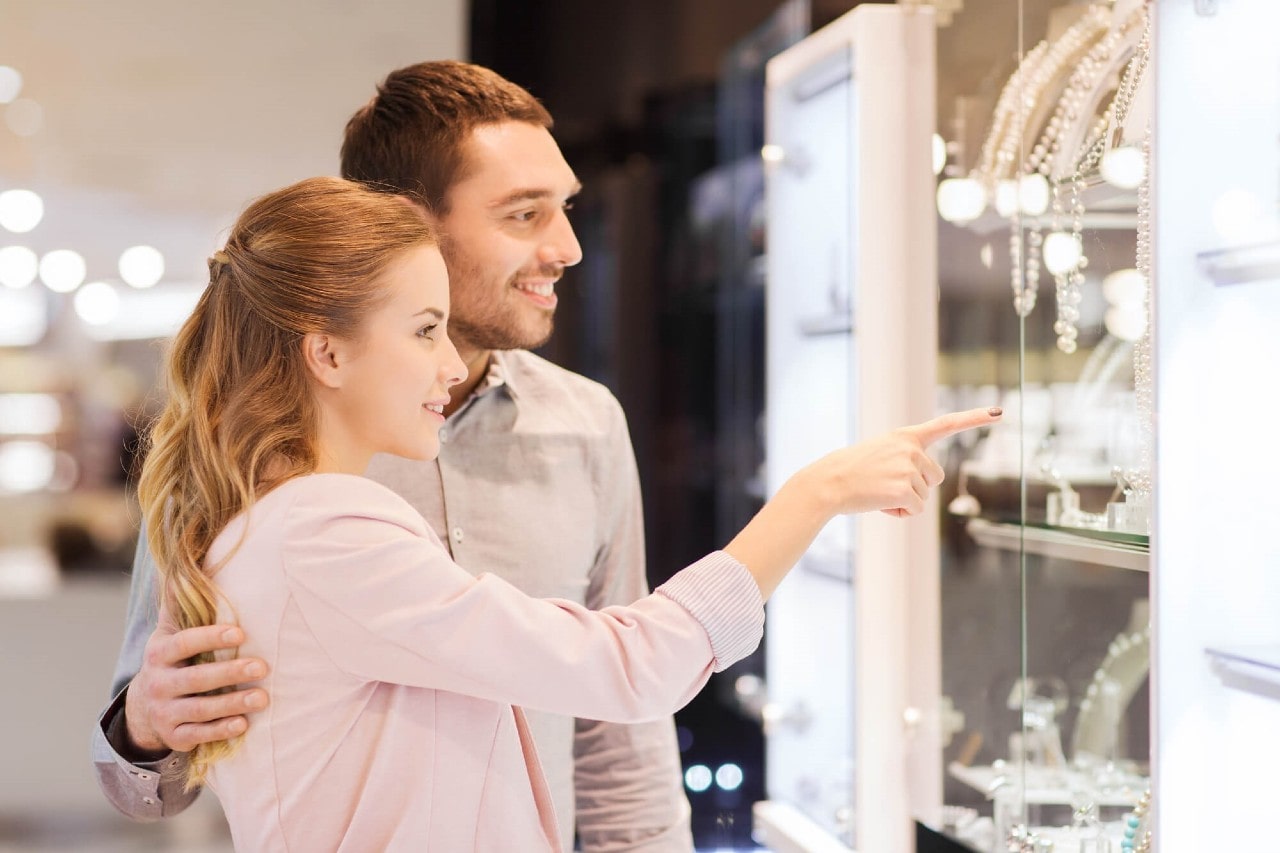 A well-dressed couple looking at a jewelry display in a brightly lit jewelry store, with the woman pointing at the jewelry.