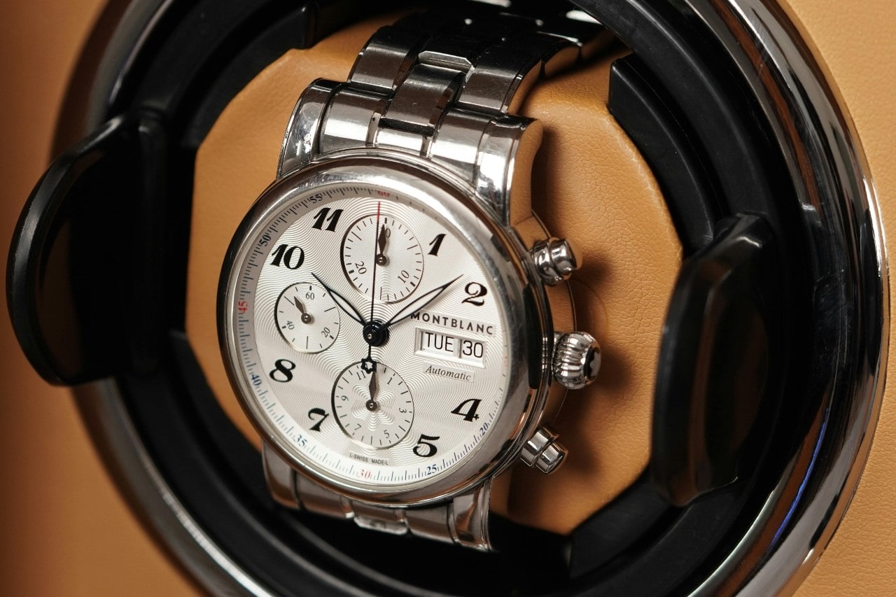 Silver Montblanc chronograph watch with a white dial, black numerals, and three subdials, displayed on a tan leather and black watch holder.