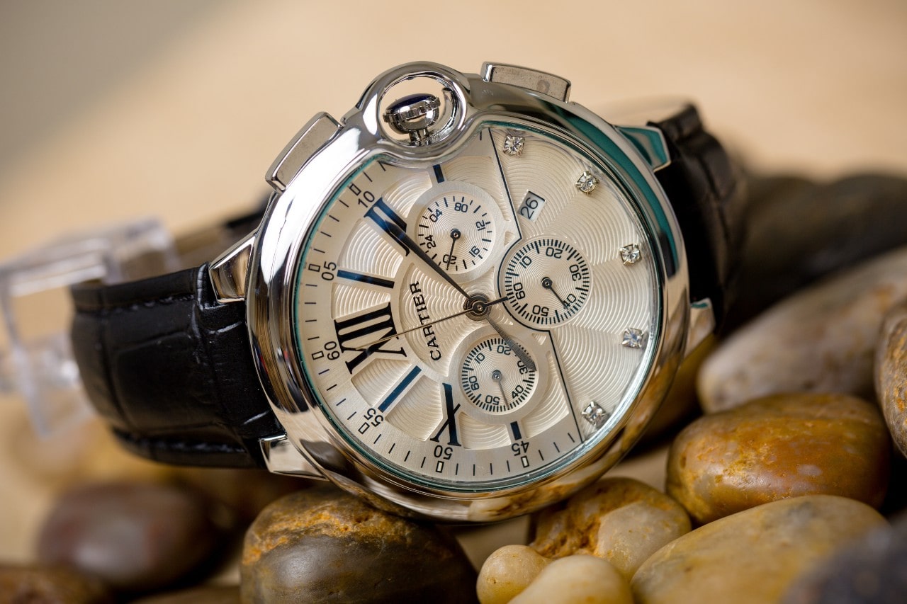 A luxurious silver wristwatch with Roman numerals and gem accents, resting on smooth pebbles.