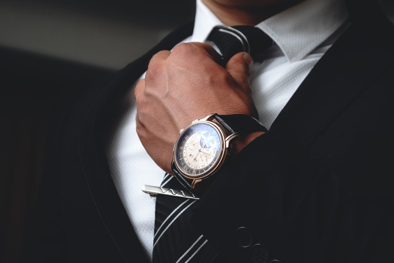 Close-up of a man in a suit adjusting his tie, wearing an elegant wristwatch with a leather strap.