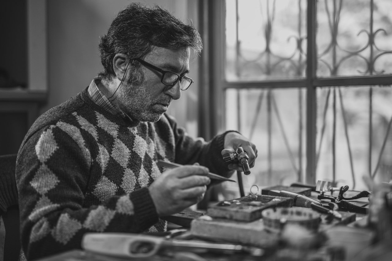 A black and white view of an artisan repairing a luxury watch in front of a large, ornate window.