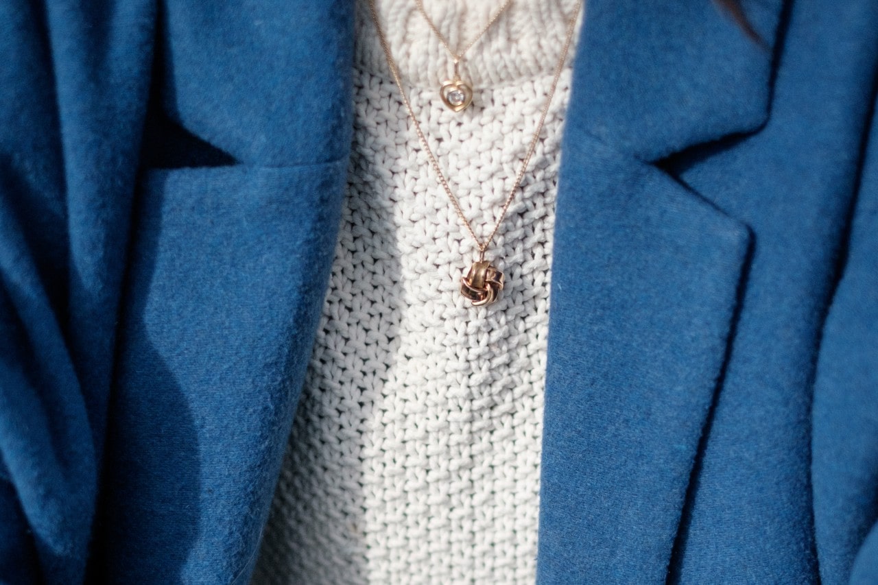 A close-up of a person wearing a wool blazer and chunky knit sweater, with two delicate gold necklaces tiered on their collarbone. A close-up of a person wearing a wool blazer and chunky knit sweater, with two delicate gold necklaces tiered on their collarbone.