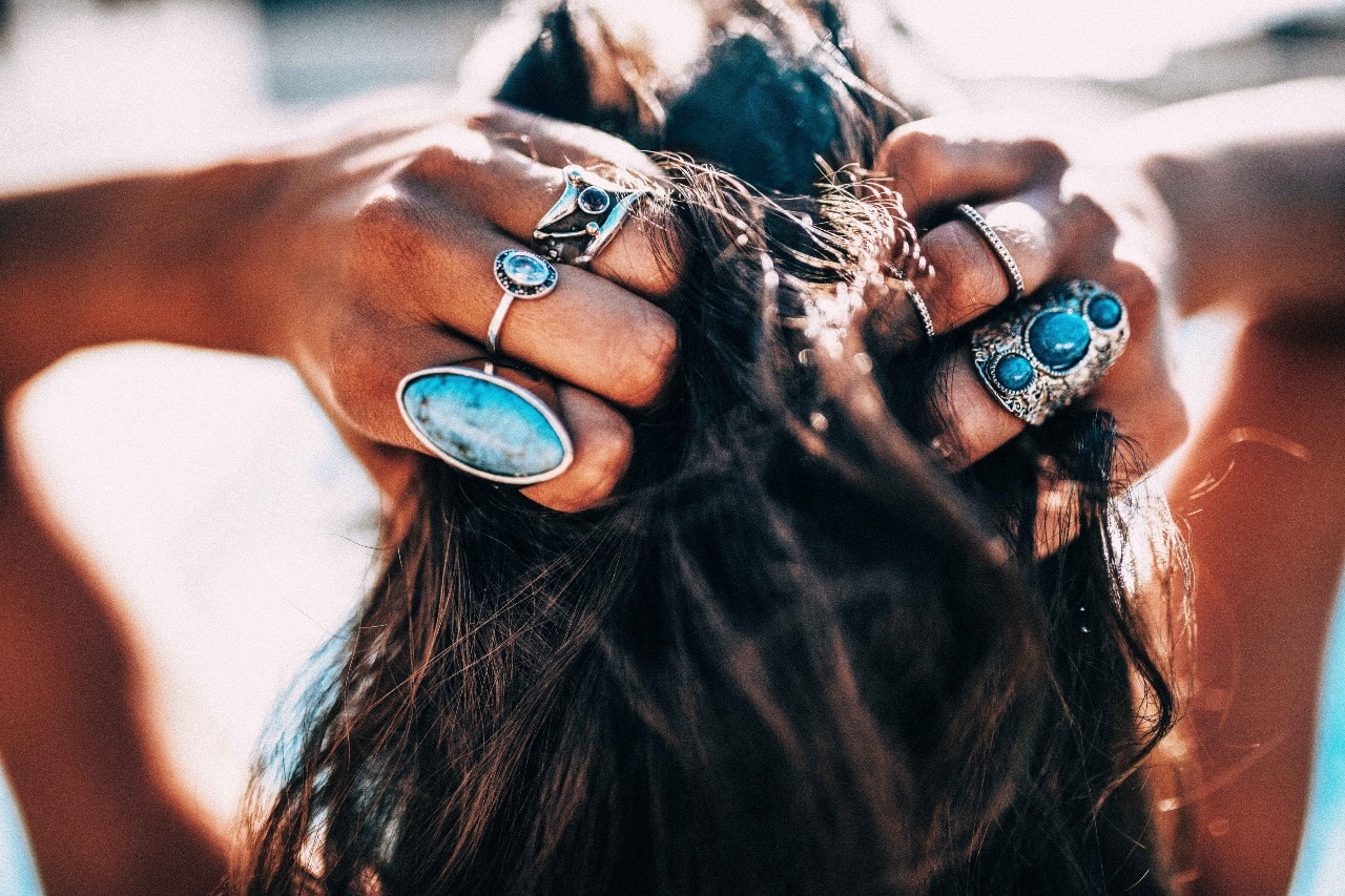 A close-up of the back of a woman’s head as she runs her fingers through her hair, her fingers adorned in multiple fashion rings. A close-up of the back of a woman’s head as she runs her fingers through her hair, her fingers adorned in multiple fashion rings.