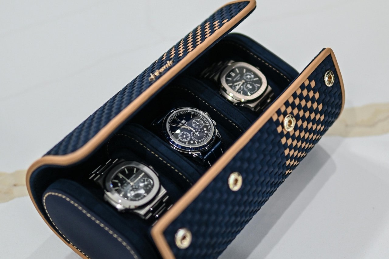 Elegant blue and tan checkered leather watch case open to reveal three luxury steel wristwatches on a light surface. Elegant blue and tan checkered leather watch case open to reveal three luxury steel wristwatches on a light surface.