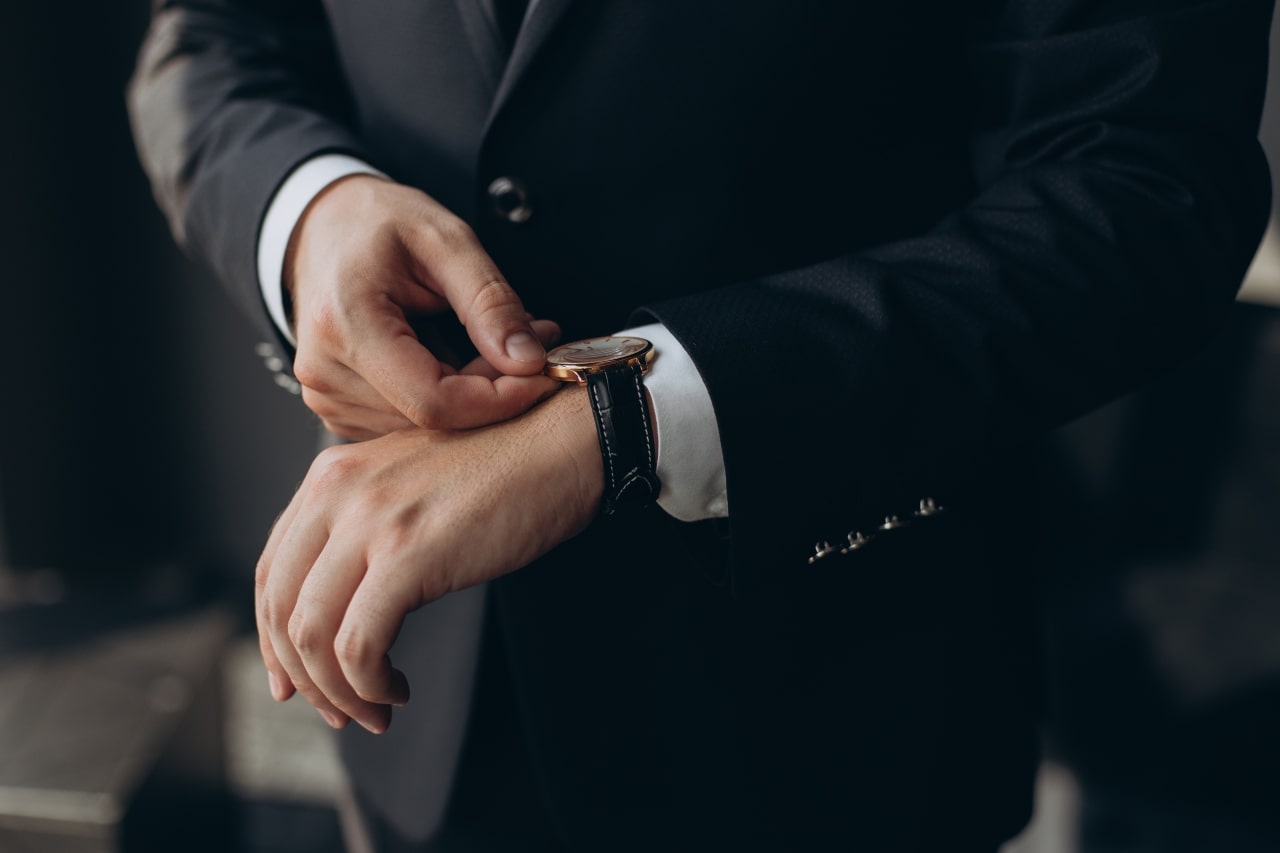 A man in a black suit fixes his wristwatch with yellow gold bezel, white dial and black leather strap. A man in a black suit fixes his wristwatch with yellow gold bezel, white dial and black leather strap.