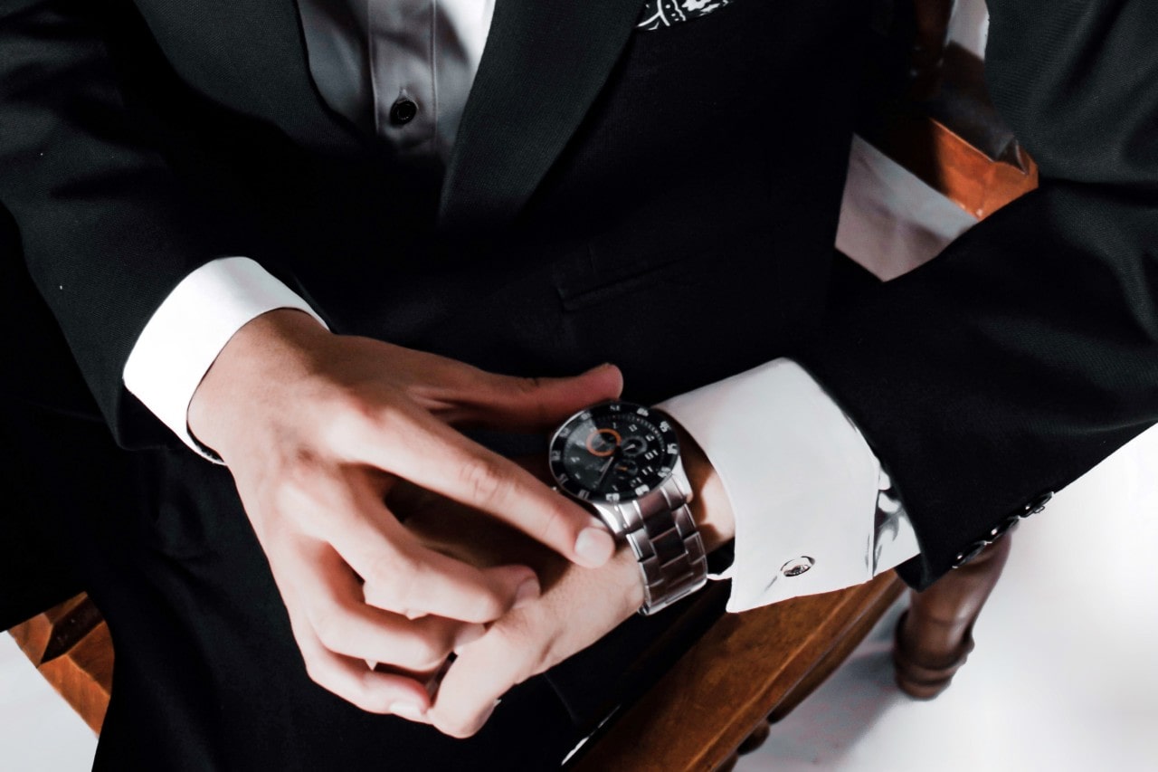 A person in a black suit adjusts a silver watch on their wrist, sitting on a wooden chair.
